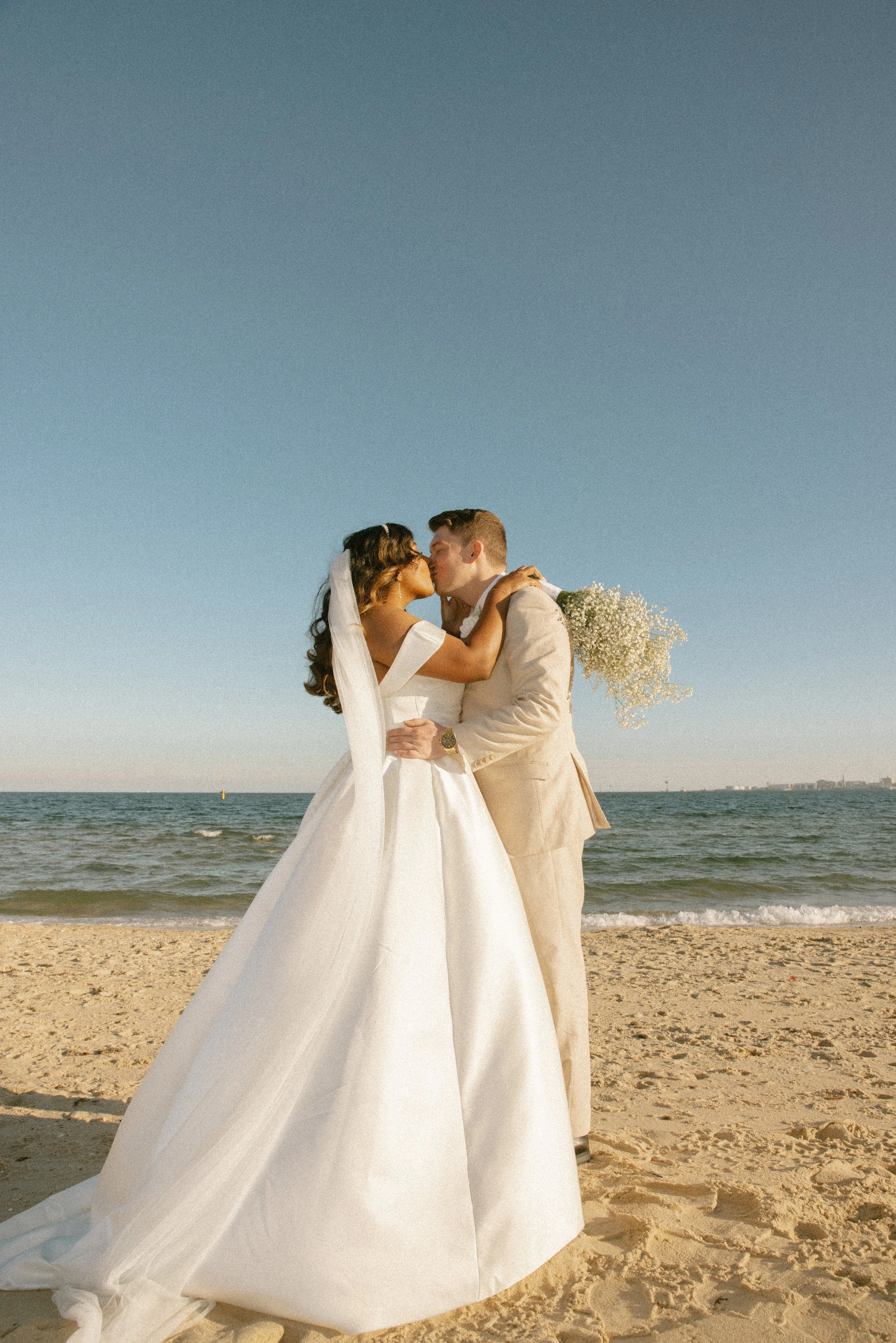 A bride and groom sharing a kiss on the beach, with the ocean and clear blue sky in the background. The bride is wearing a white wedding dress and veil, while the groom is in a beige suit and holding a bouquet of small white flowers.