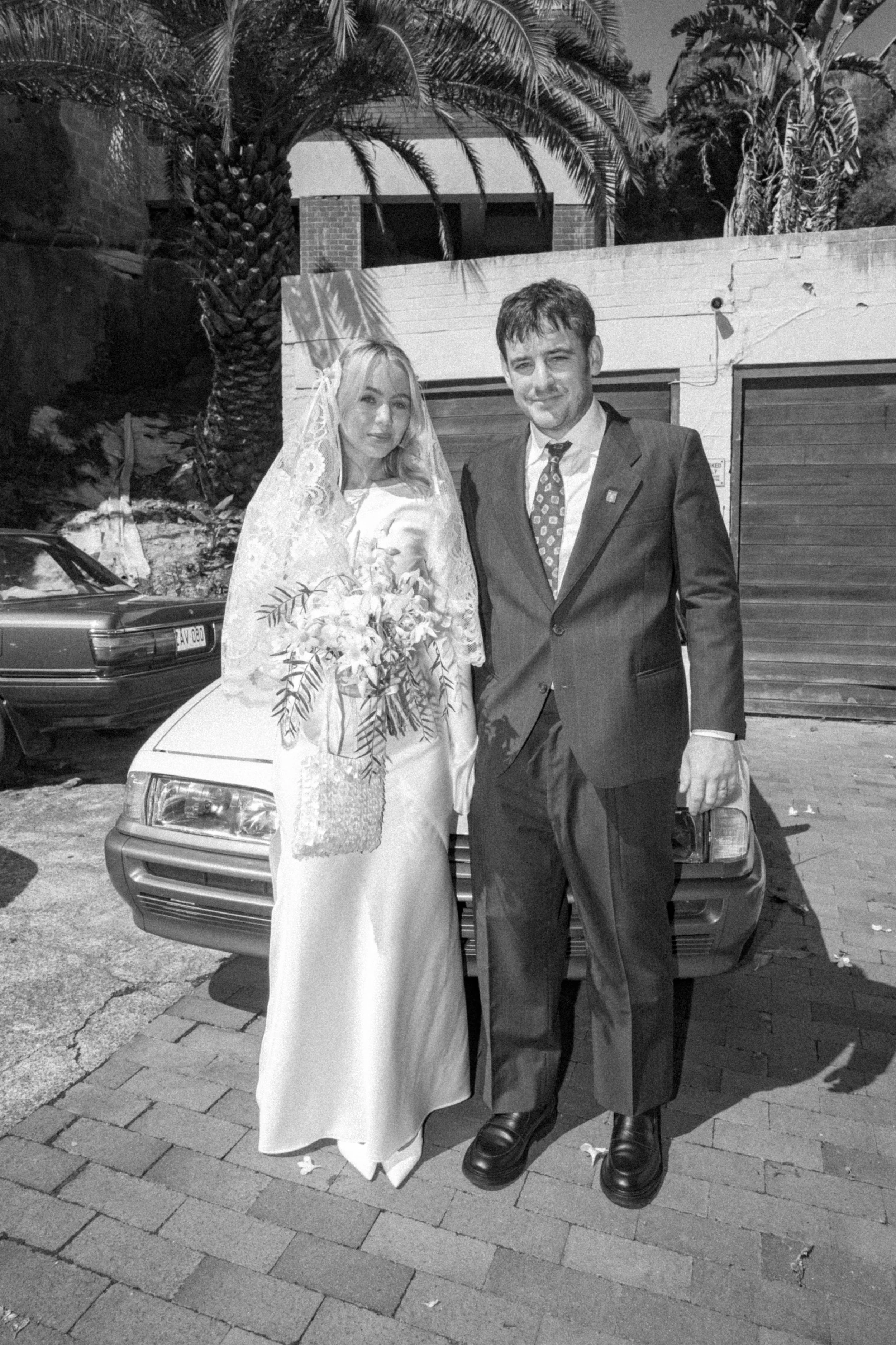 A bride and groom standing in front of a car, with palm trees and a building in the background. The bride is wearing a wedding dress and veil, holding a bouquet, and the groom is in a suit with a patterned tie.