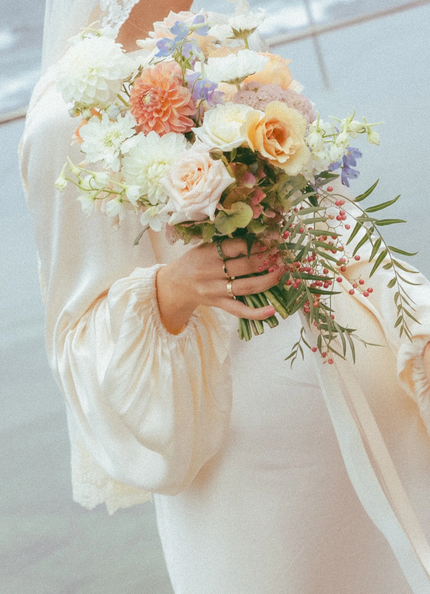 Person holding a bouquet of pastel-colored flowers, including white, peach, purple, and pink blossoms, against a softly textured background.