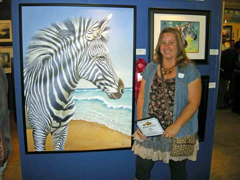 A woman standing next to a large framed painting of a zebra on a beach, holding an award certificate at an art exhibit in San Diego.