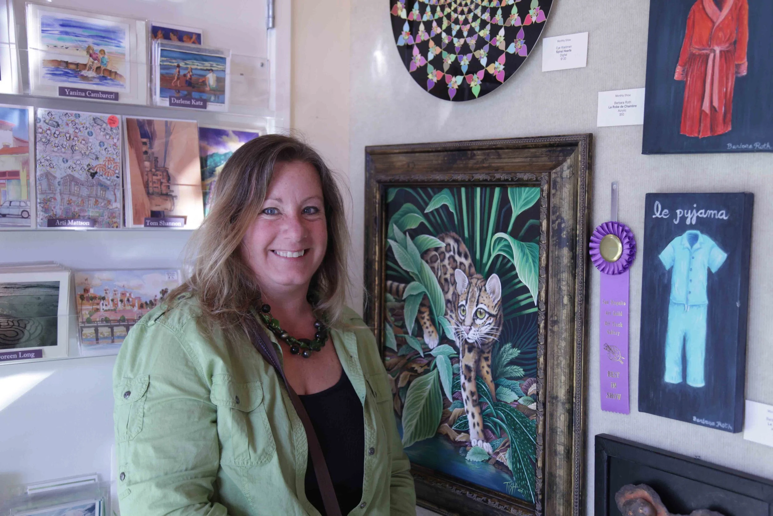 A woman with shoulder-length wavy hair, wearing a light green jacket and a black necklace, standing beside a framed painting of a ocelot in a jungle setting, at an art gallery with various artworks displayed on the wall.