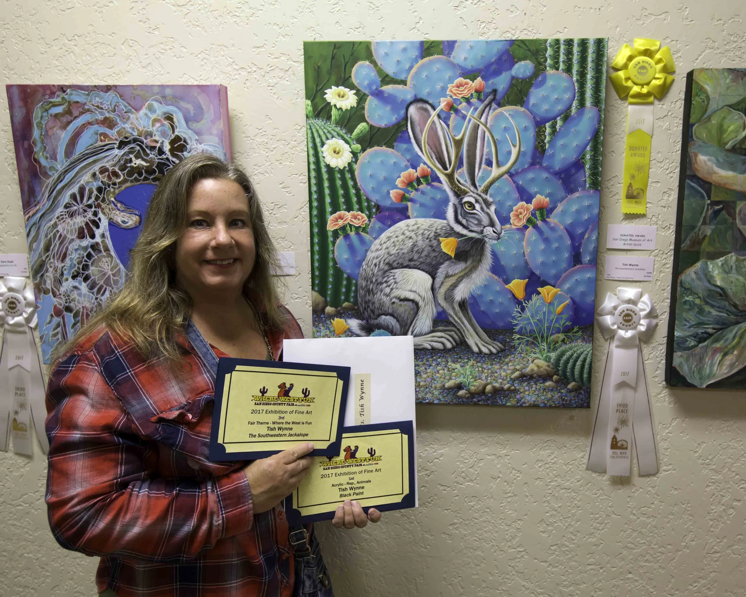 Woman in plaid shirt holding award certificates, standing next to a colorful painting of a jackrabbit with tall cacti and blooming flowers, with ribbons on the wall behind.