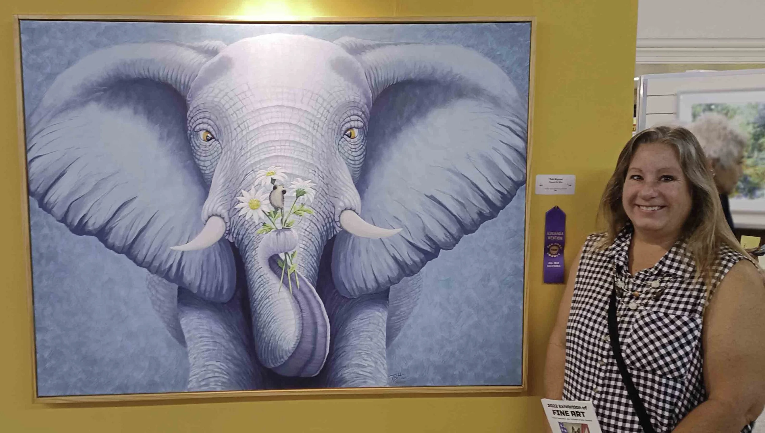 A woman standing next to a framed painting of an elephant with flowers and a mouse on its trunk, smiling at an art exhibition.
