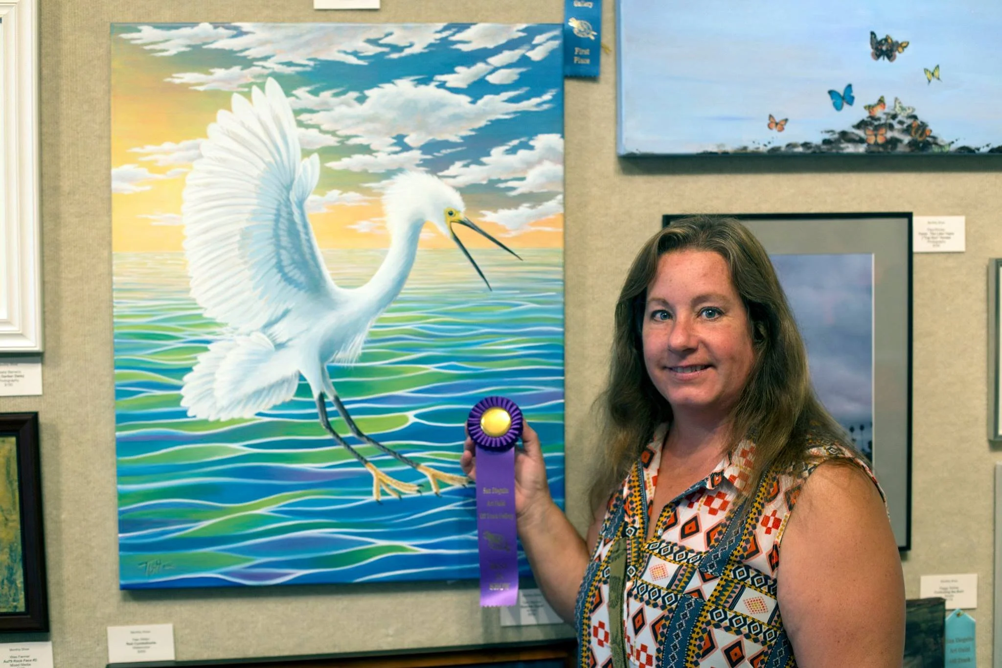 A woman with long brown hair wearing a sleeveless patterned top holding an award ribbon stands next to a painting of a white egret flying over water with a sunset sky in the background.