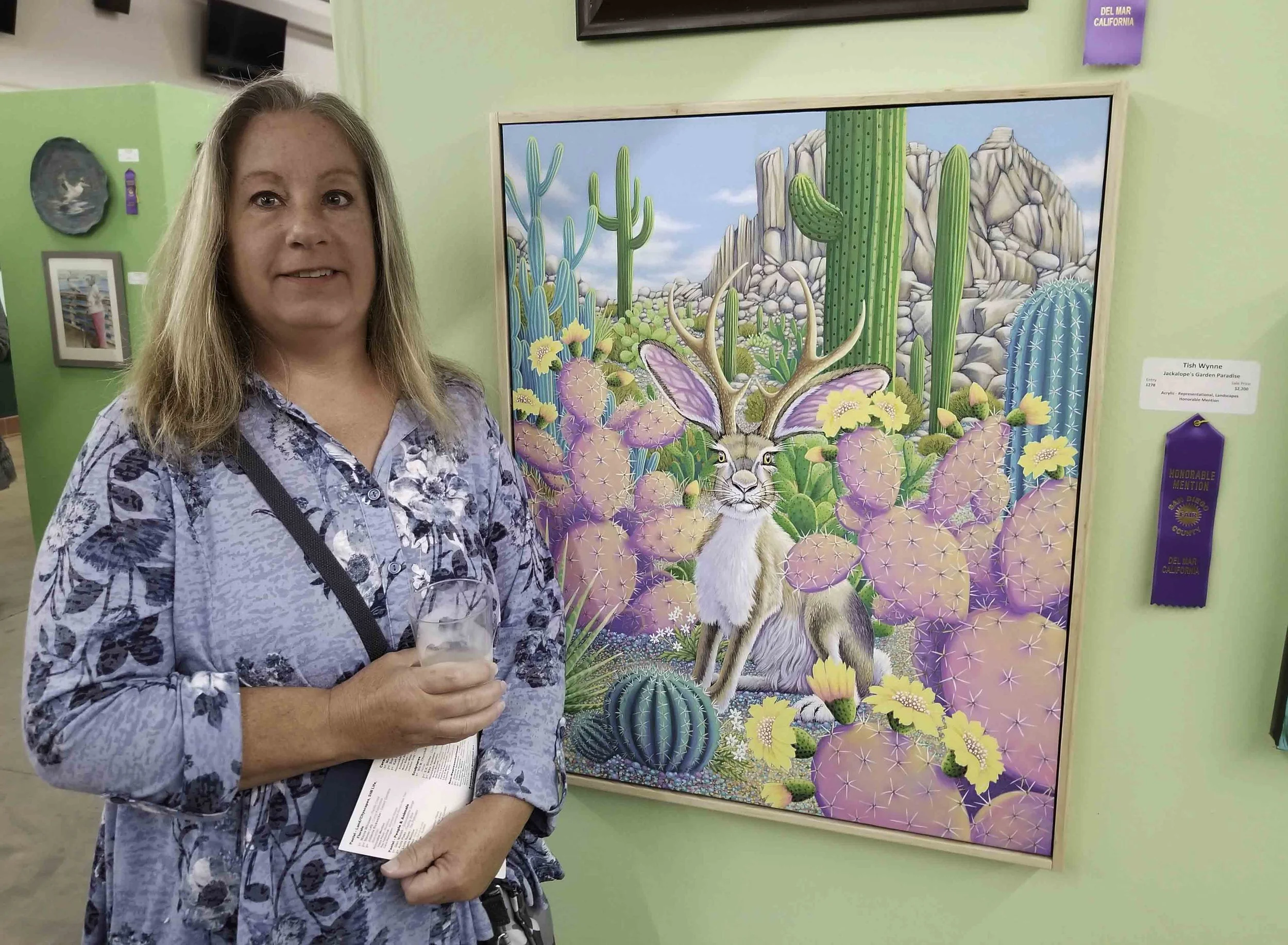 A woman with blonde hair holding a pamphlet, standing next to a colorful desert-themed painting of a jackalope and cacti in an art gallery.