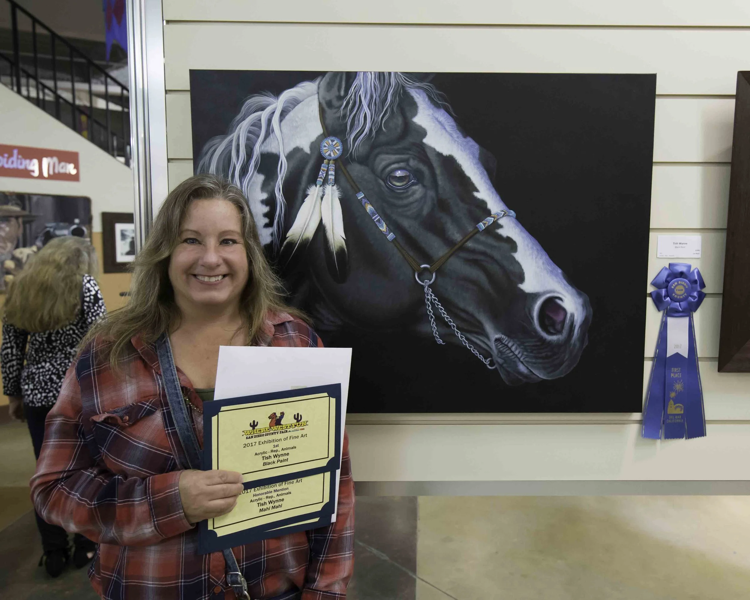 Woman holding award certificates standing in front of a painted portrait of a horse with a blue ribbon on its bridle at an art exhibition.