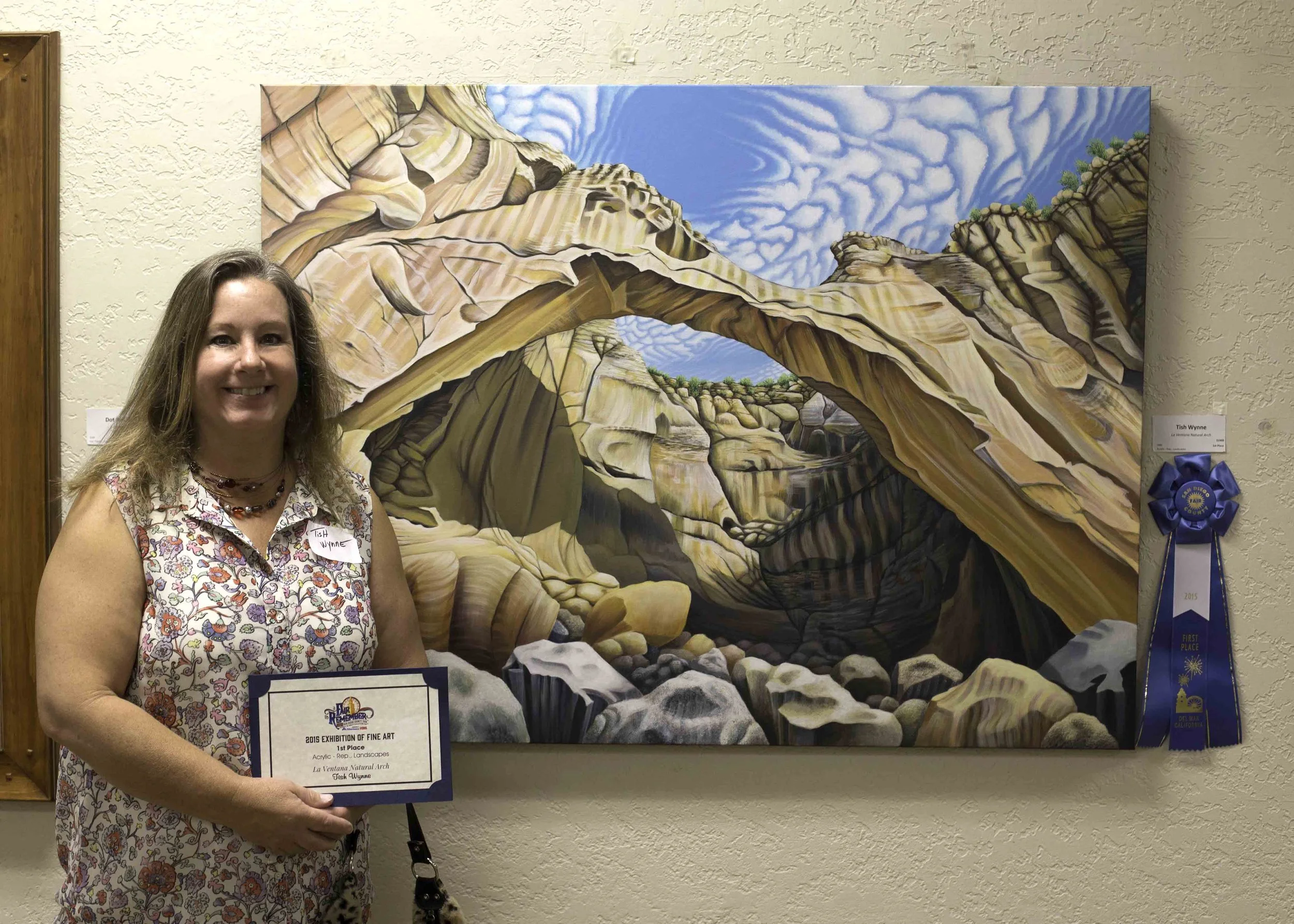 A woman standing next to a colorful painting of a desert canyon with rock formations, trees, and blue sky with clouds, holding a certificate of achievement with a blue ribbon on the wall beside her.