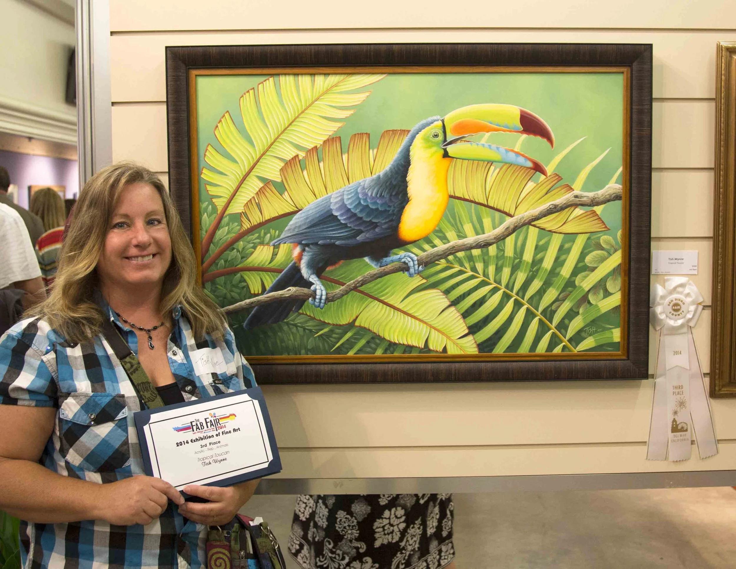 A woman holding an award plaque standing next to a colorful painting of a toucan with a large yellow, orange, green, and red beak, perched on a branch surrounded by jungle green leaves.