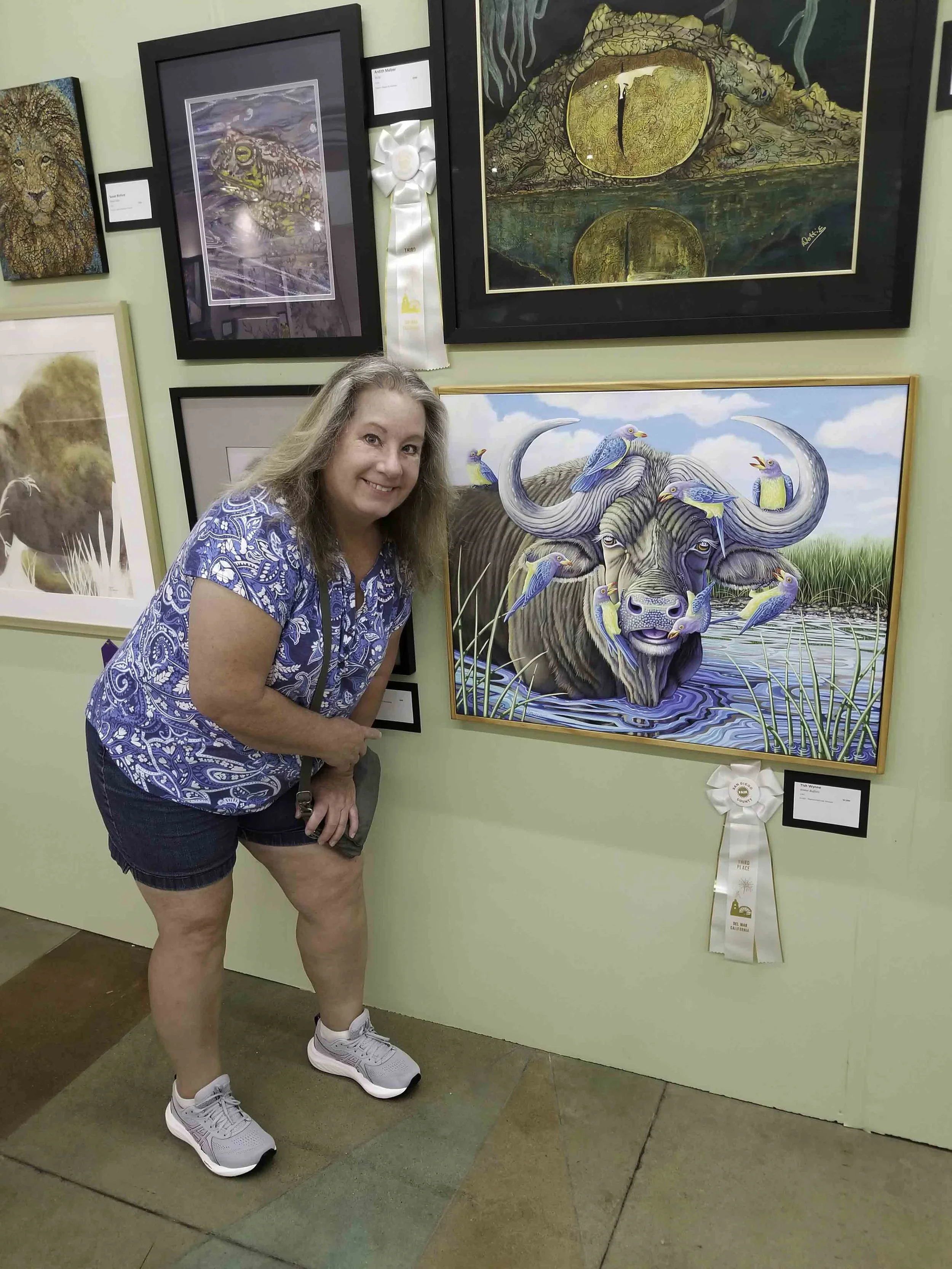 A woman in a blue patterned shirt and shorts, smiling and posing in front of her artwork at an art gallery. The artwork features a water buffalo with blue birds perched on and around it, set in a water landscape.