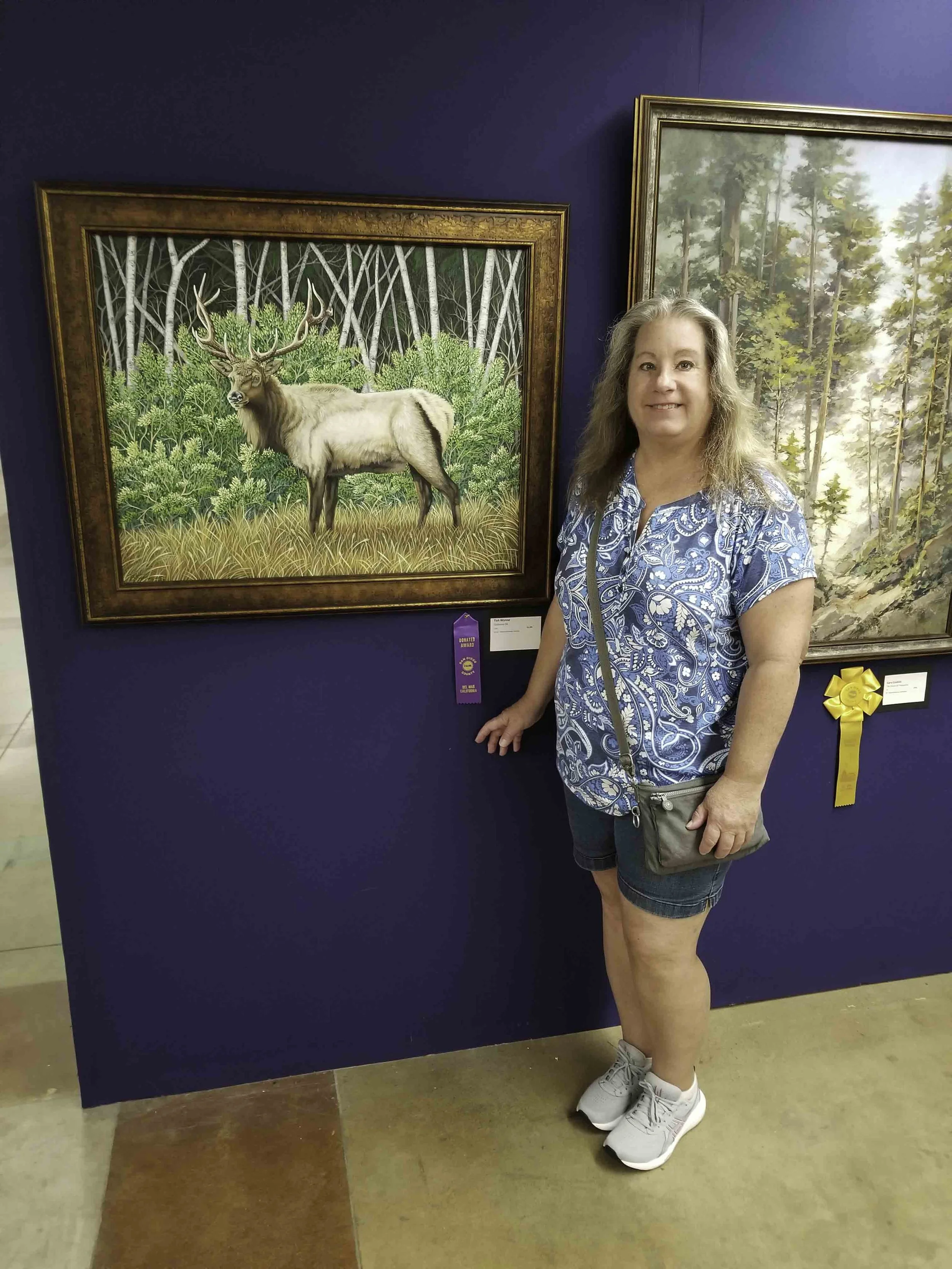 Woman standing next to a painting of a male elk in a forest at an art show.