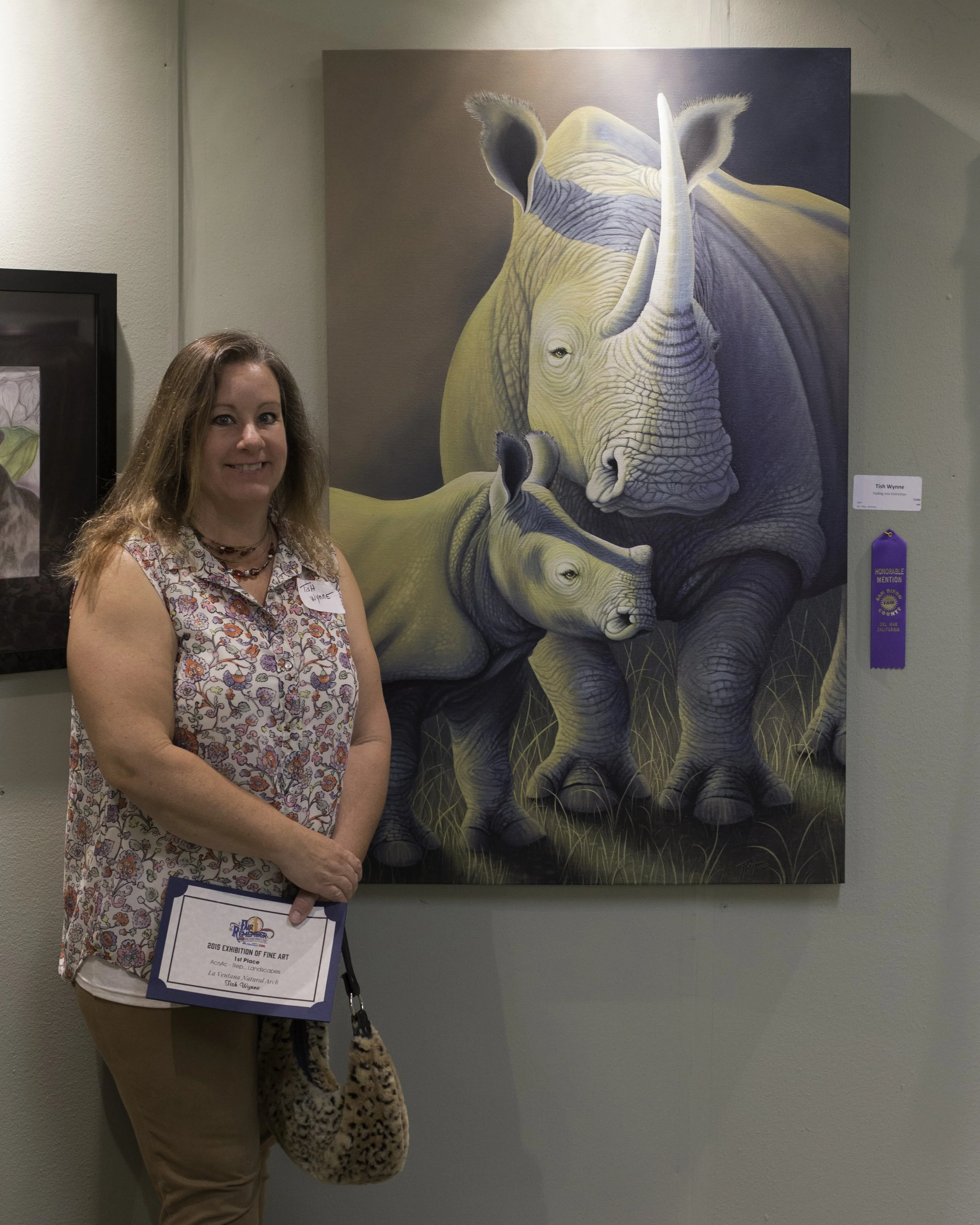 Woman standing in front of a painting of two rhinoceroses, one large and a baby rhino, on a gallery wall. She is holding a certificate award.