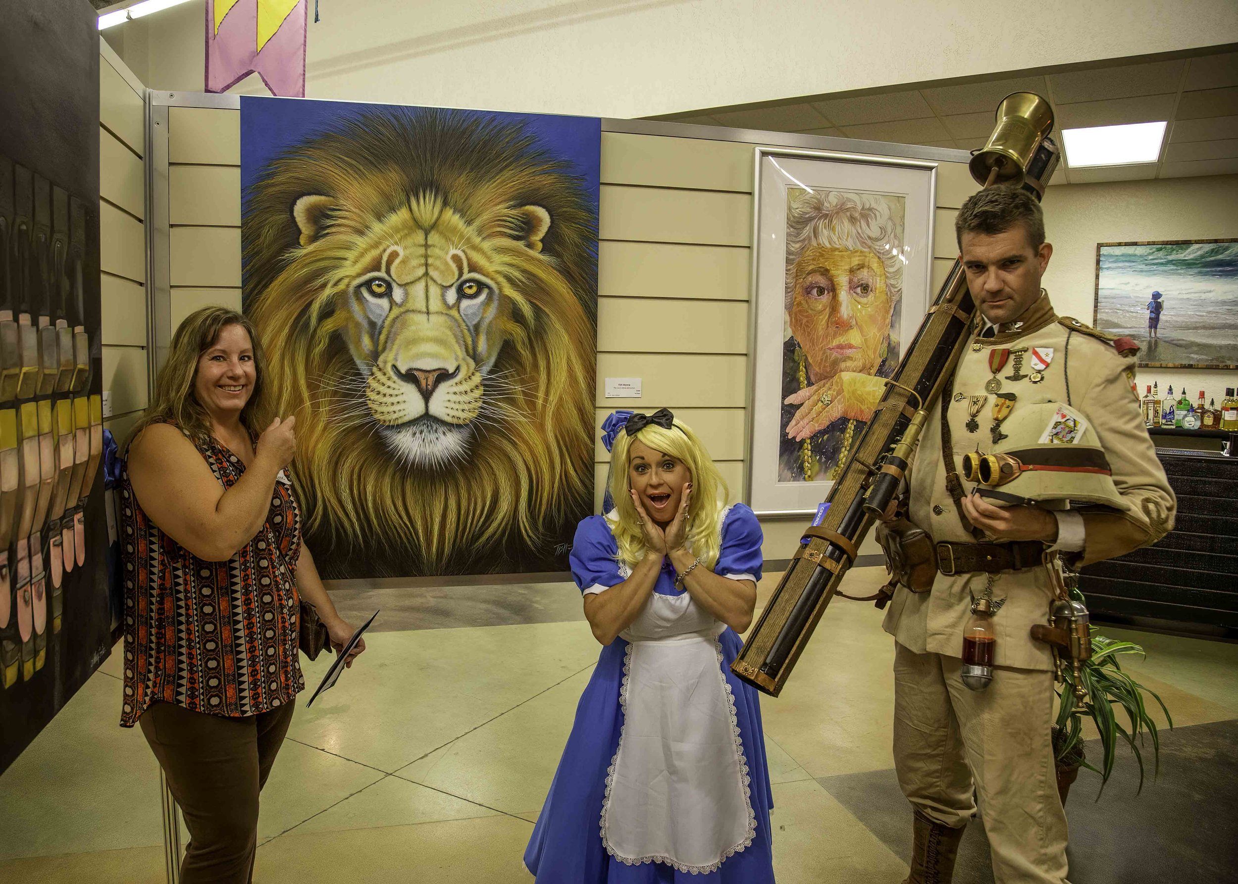women and a man in costume at art exhibit. The woman on the left is smiling and pointing at a large painting of a lion. The woman in center is dressed as Alice from Alice in Wonderland, with surprised expression. The man on right is dressed as a sold