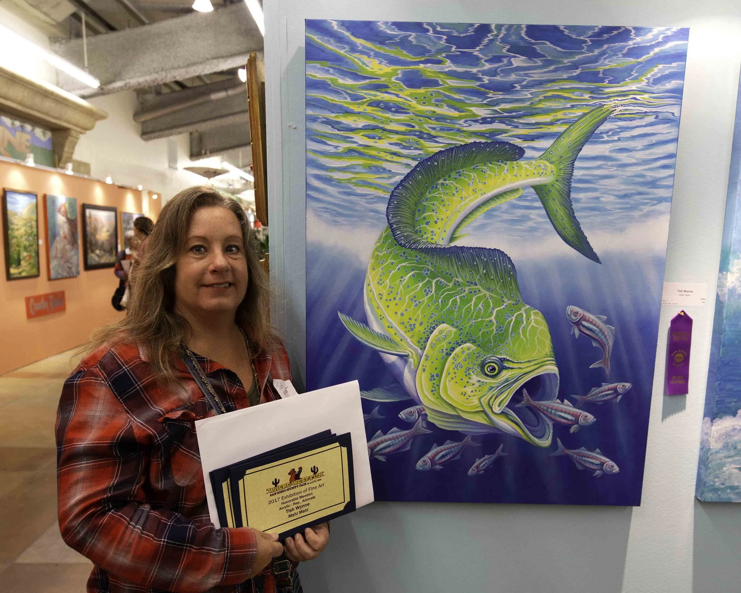 A woman with shoulder-length brown hair wearing a red plaid shirt holding a certificate standing next to a colorful painting of a large fish underwater with smaller fish surrounding it at an art gallery.