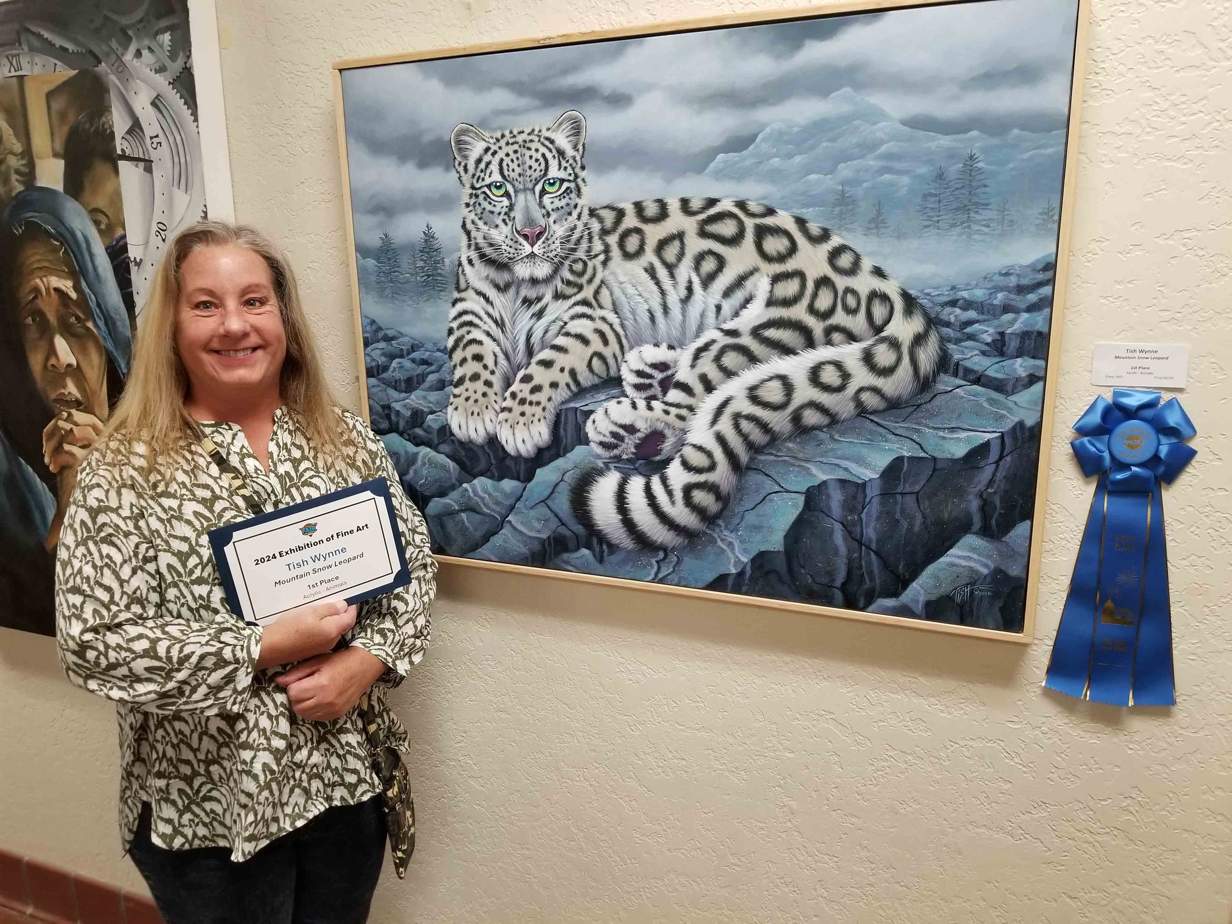 Woman smiling and holding a certificate in front of a painting of a snow leopard lying on rocks with a mountain landscape in the background, and a blue ribbon award is displayed on the wall.