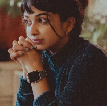 A young woman with short curly hair looking thoughtful, resting her chin on her folded hands, wearing a dark sweater and a smartwatch.