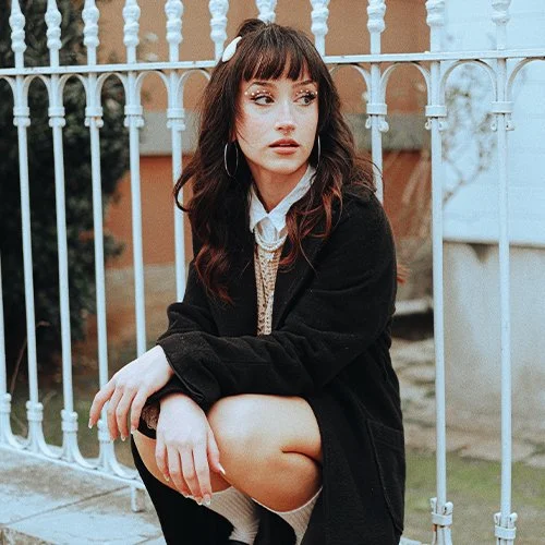 Young woman with dark hair and bangs, wearing a black jacket, sitting by a white fence outdoors.