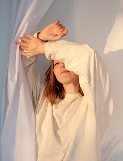 A woman with light brown hair wearing a white shirt, raising her left arm with her hand near her face, standing among white curtains or fabric.