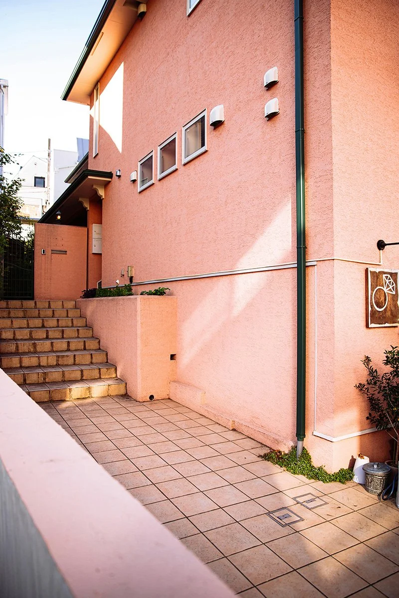 A pink stucco house with stairs leading up to a door, small square windows, and exterior wall-mounted lights. There are steps and a small patio area at the front.