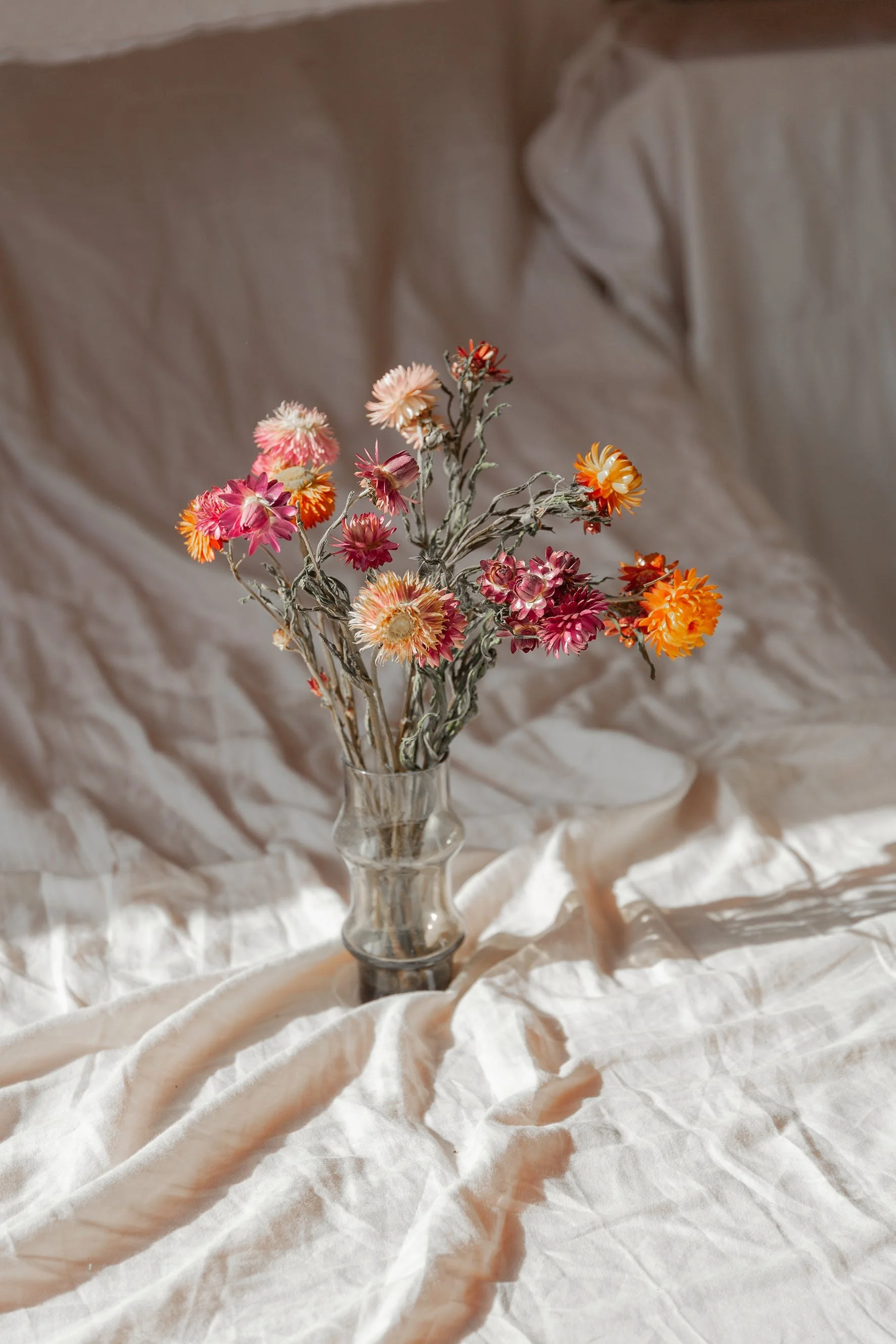 A clear glass vase with a bouquet of dried flowers, including pink, orange, and cream-colored blooms, set against a crumpled beige fabric backdrop.