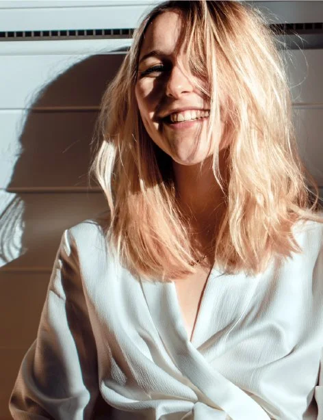 A woman with blonde, wavy hair smiling, wearing a white blouse, with sunlight casting shadows on her face and background.