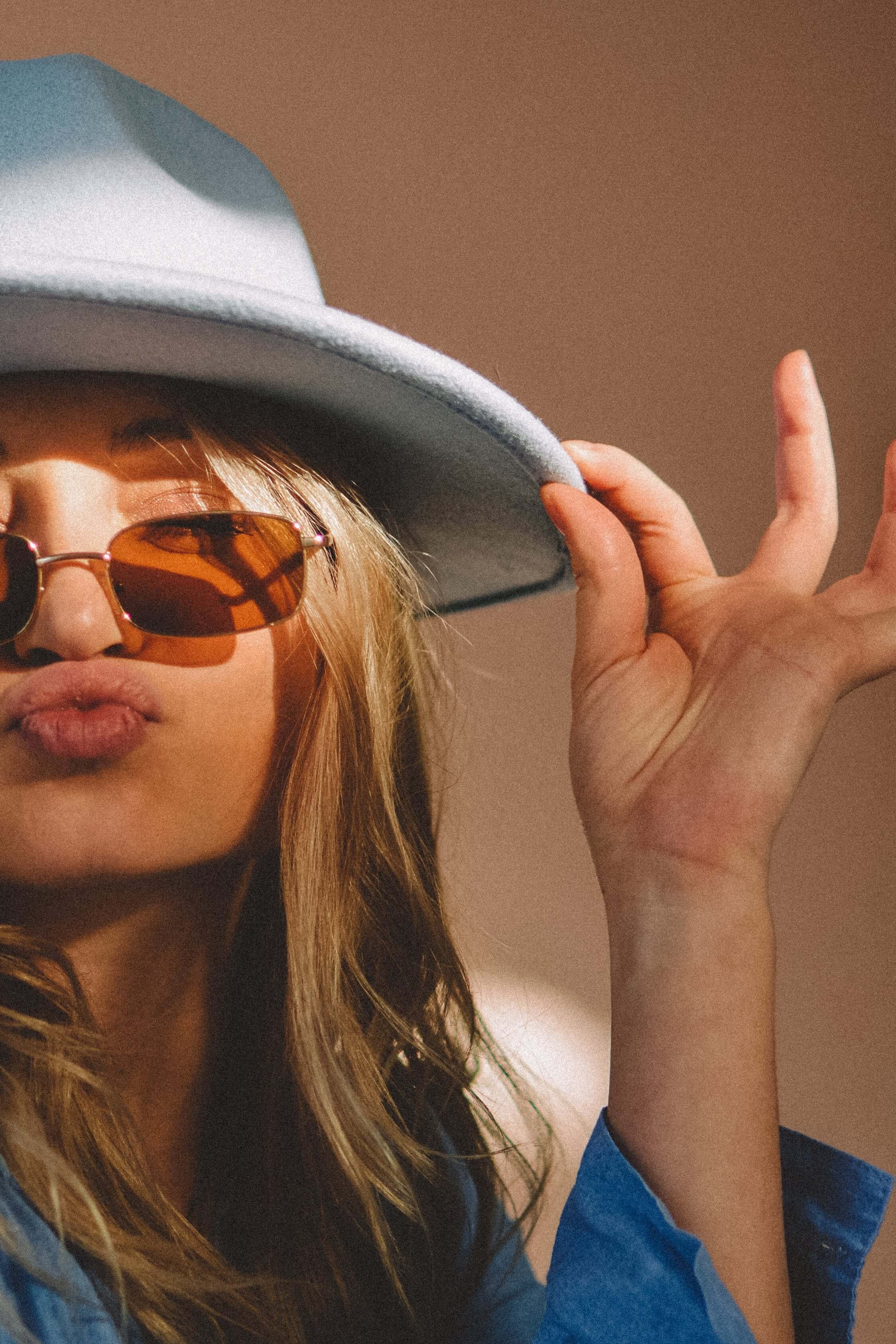Close-up of a woman wearing a gray hat, brown sunglasses, and a blue jacket, making a playful face with her lips puckered. She has long, wavy blonde hair and is holding the brim of her hat with her right hand.