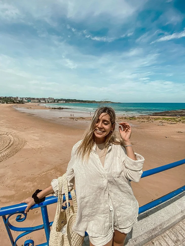 Smiling young woman in beige outfit standing by a blue railing on a beach with sandy shore, rocky areas, and ocean waves under a partly cloudy sky