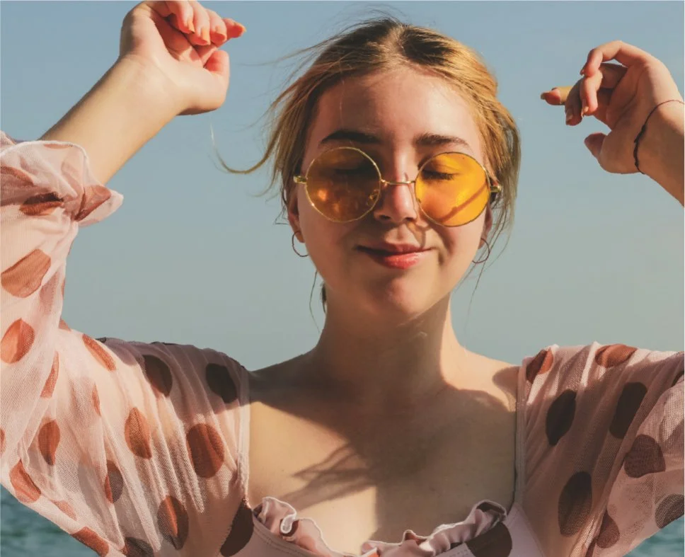 A young woman with blonde hair wearing yellow-tinted sunglasses and a pink top with polka dots, smiling with eyes closed, outdoors on a sunny day.