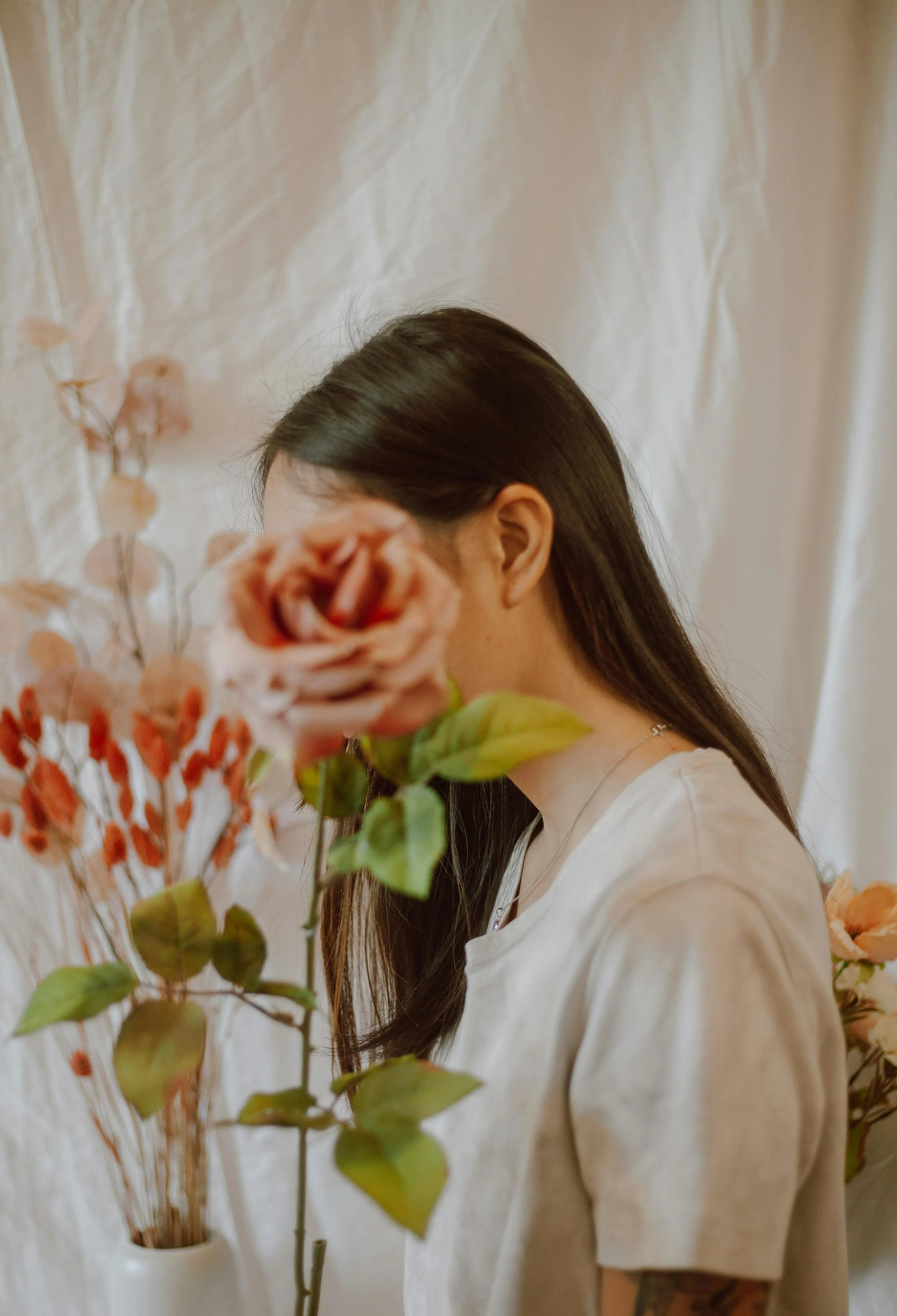 A woman with long dark hair is holding a bouquet of pink and orange roses in front of her face, obscuring her facial features, against a light-colored curtain background.