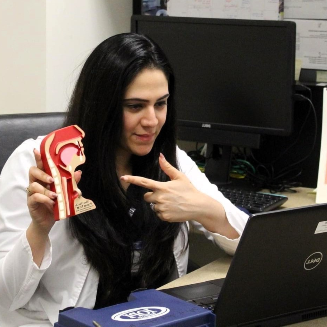 Rinki Varindani Desai wears a white lab coat sitting at a desk, holding a model of a human ear and pointing at her own ear while looking at her laptop.