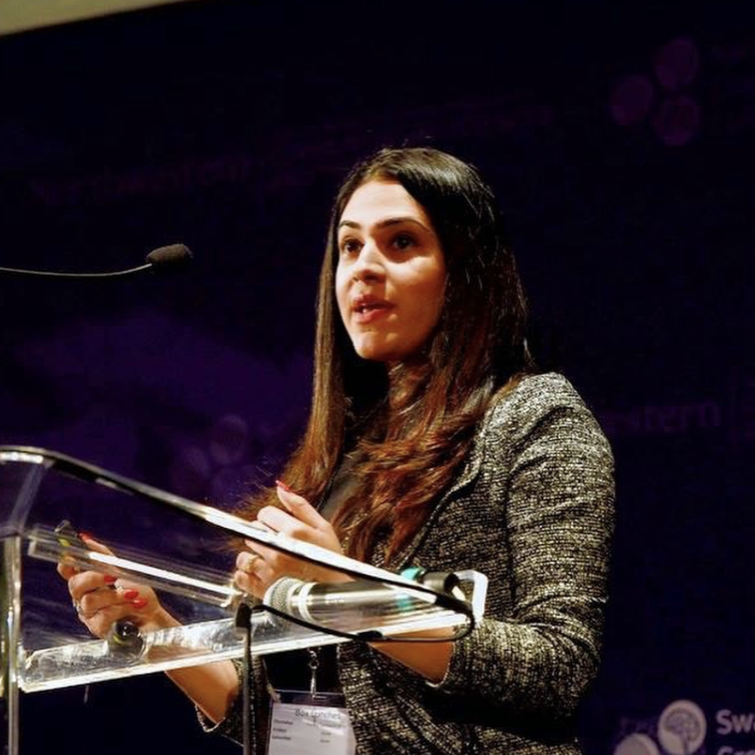 A woman with long dark hair standing behind a glass podium, speaking at an event, with a dark purple background.
