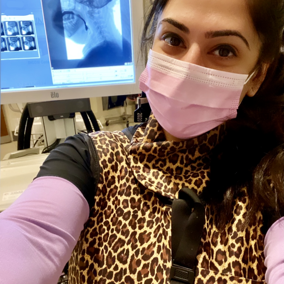 Rinki Varindani Desai wearing a pink face mask and leopard print clothing taking a selfie in a medical setting with an X-ray monitor in the background.