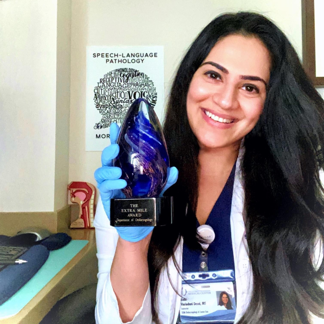 Rinki Varindani Desai wearing a white coat and blue gloves, holding a glass award shaped like a blue flame with a black base that reads 'The Extra Mile Award, Department of Otolaryngology.'