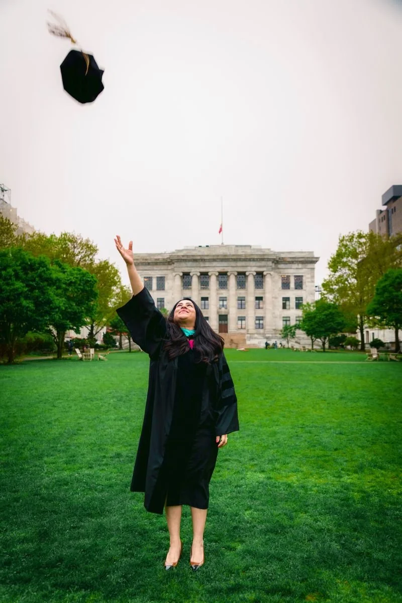 Rinki Varindani Desai in a graduation cap and gown standing on a grassy area in front of a government building, throwing her cap into the air and smiling.
