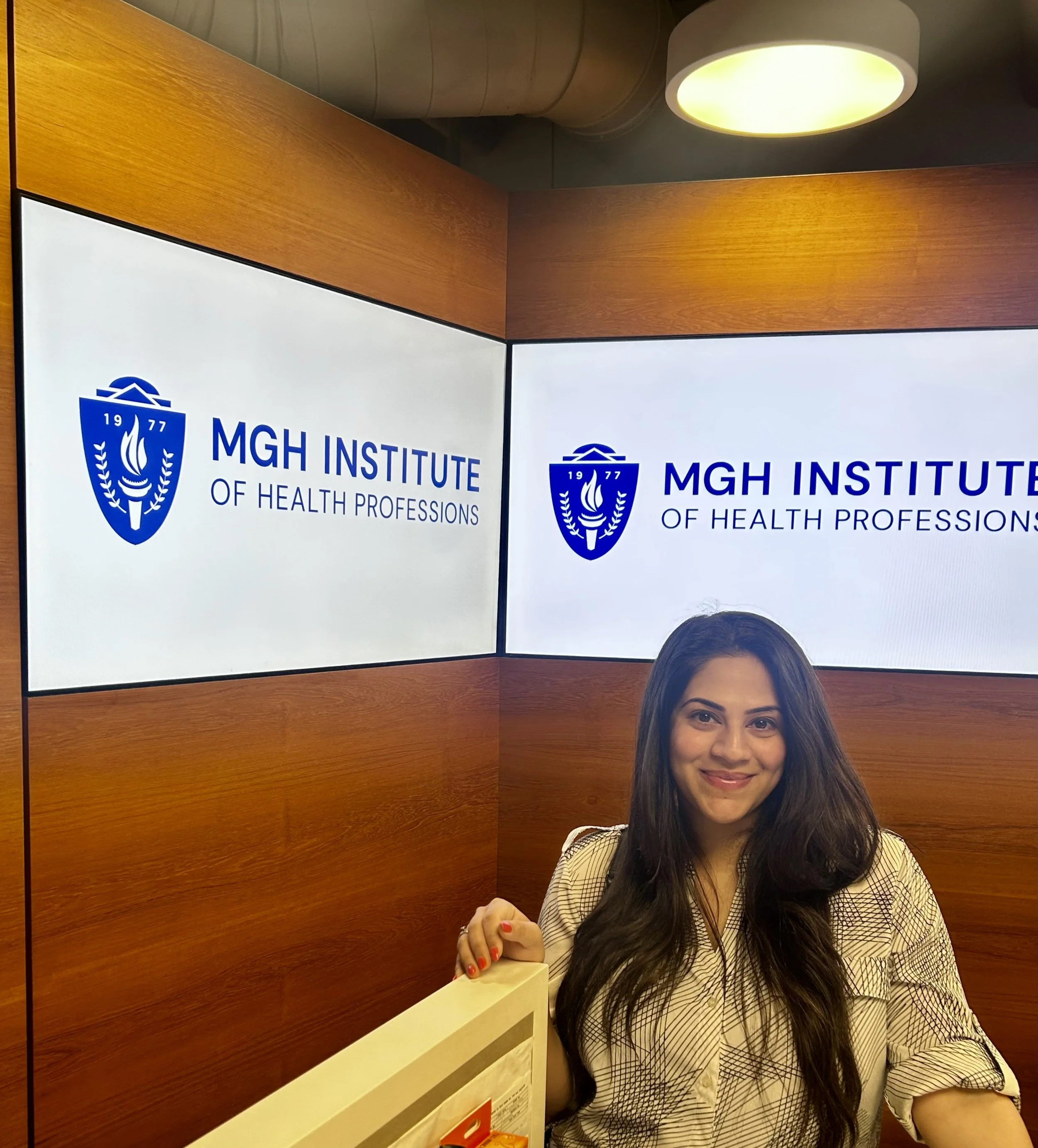 Rinki Varindani Desai, smiling, sitting in an elevator, wearing a beige and black striped blazer, with a wooden wall and digital screen displaying the 'MGH Institute of Health Professions' logo behind her.