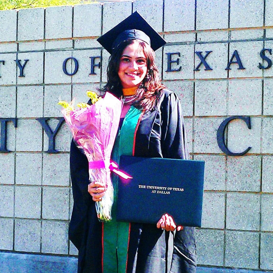 Rinki Varindani Desai in a graduation gown and cap holding a bouquet of flowers and a diploma in front of a stone wall that has the text 'University of Texas at Dallas' on it.