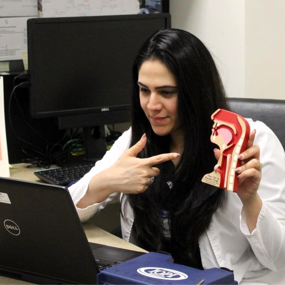 Rinki Varindani Desai, wearing a white lab coat, pointing at a model of a human ear, with a computer and monitors in the background, in an office or medical setting.