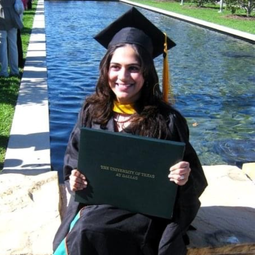 Rinki Varindani Desai in graduation attire, including a cap and gown, sitting by a water feature and holding a diploma.