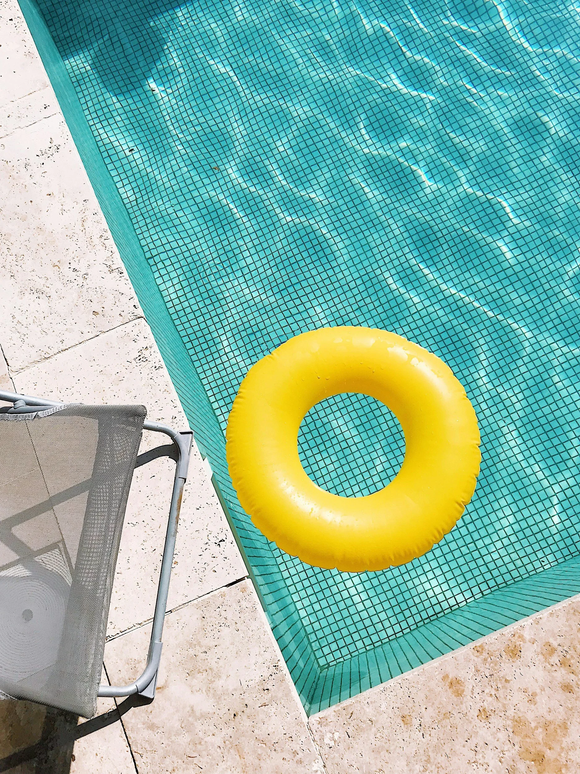 A swimming pool with blue mosaic tiles, a yellow inflatable ring floating on the water, a lounge chair with a mesh surface, and a tan stone deck area.