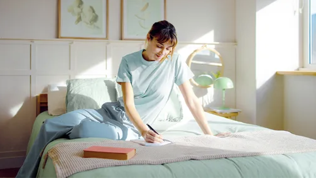 A woman sits on her bed, journaling in her notebook.