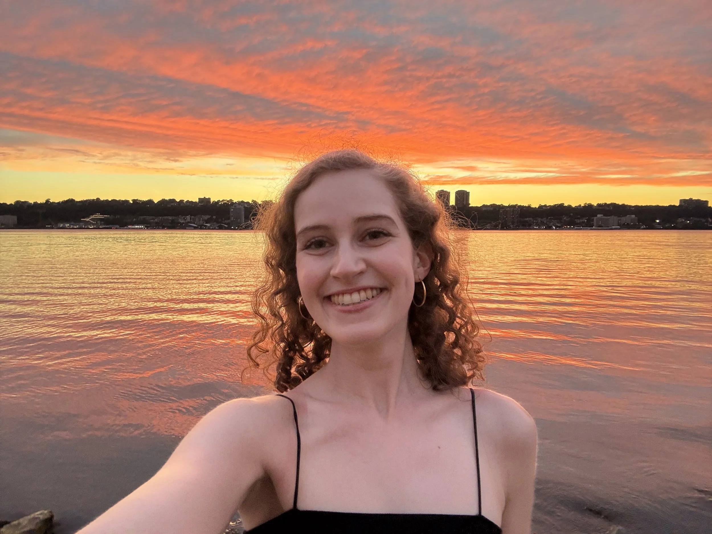 A woman with curly brown hair smiles in front of an orange and yellow sunset reflecting on the river.