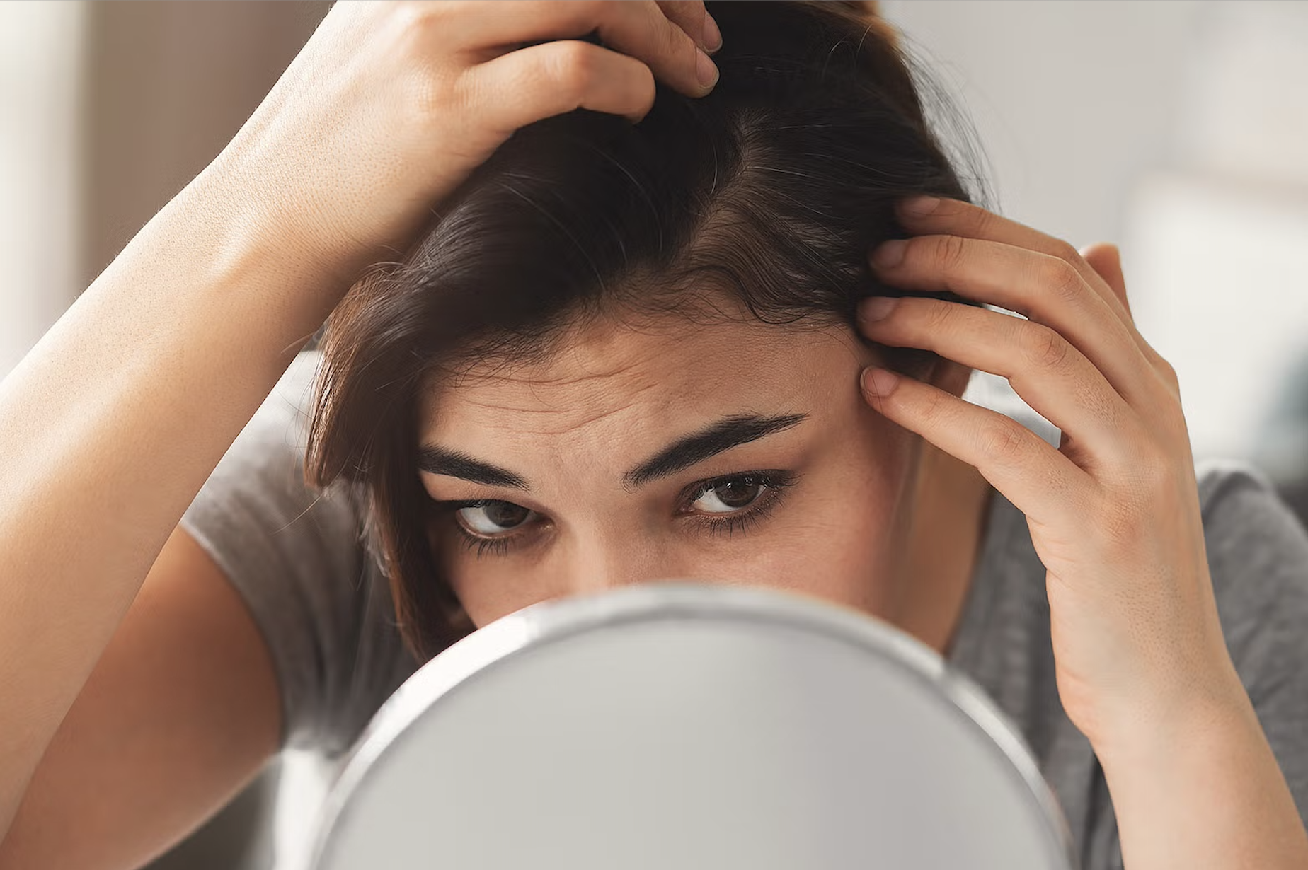A woman gazes into a mirror, her hands on her scalp.