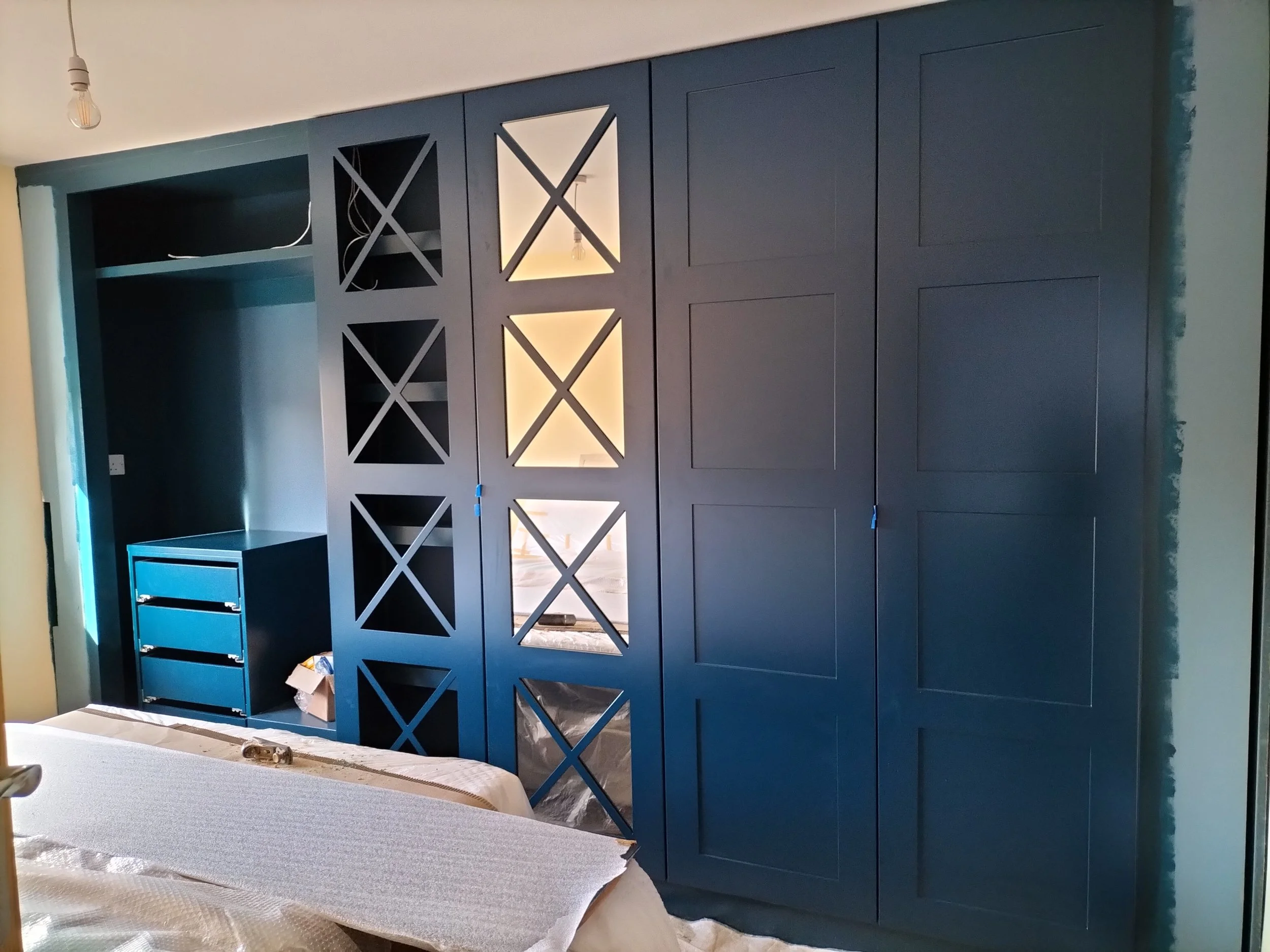 Blue built-in wardrobe with closed cabinets, open shelves, and decorative lattice panels under construction in a bedroom.