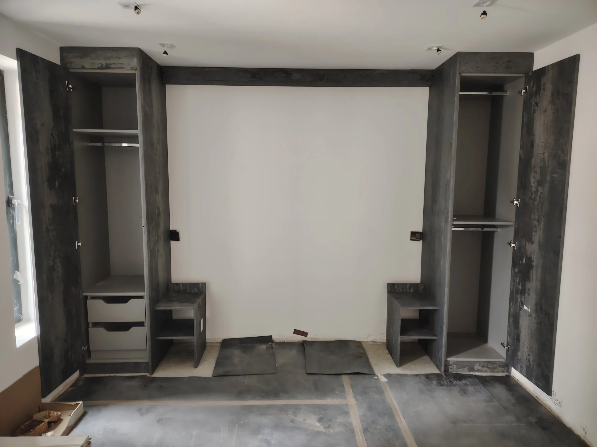 Unfinished built-in closets with open doors in a room under construction, featuring a white wall and partially installed flooring.