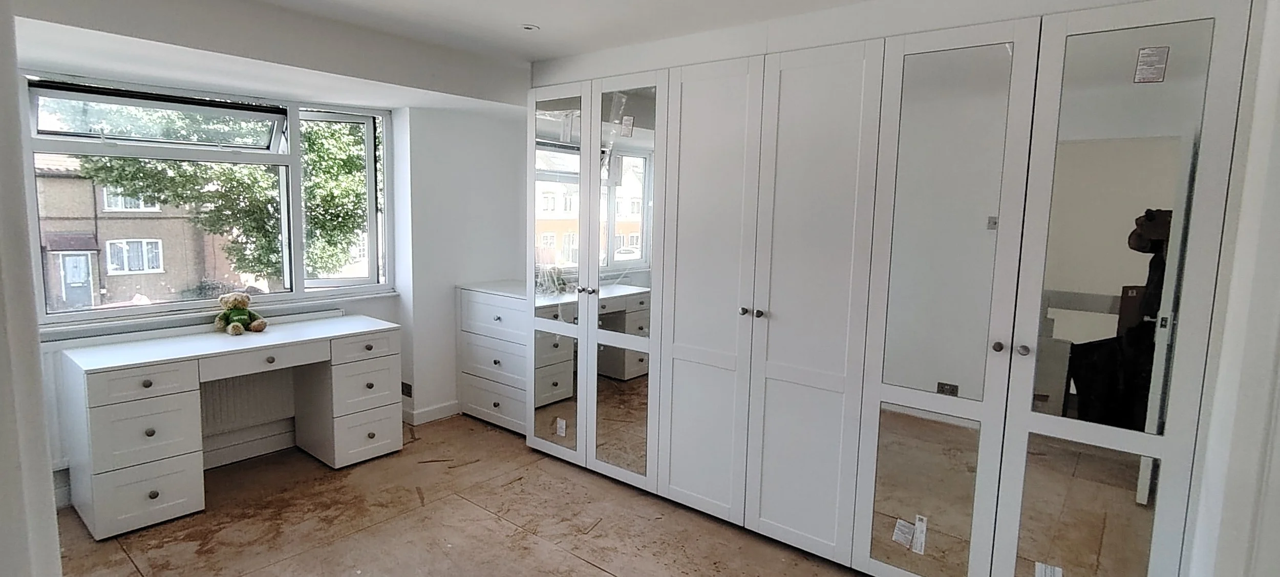 Empty room with white built-in cabinets, a white desk, and a window with a view of trees and neighboring buildings.
