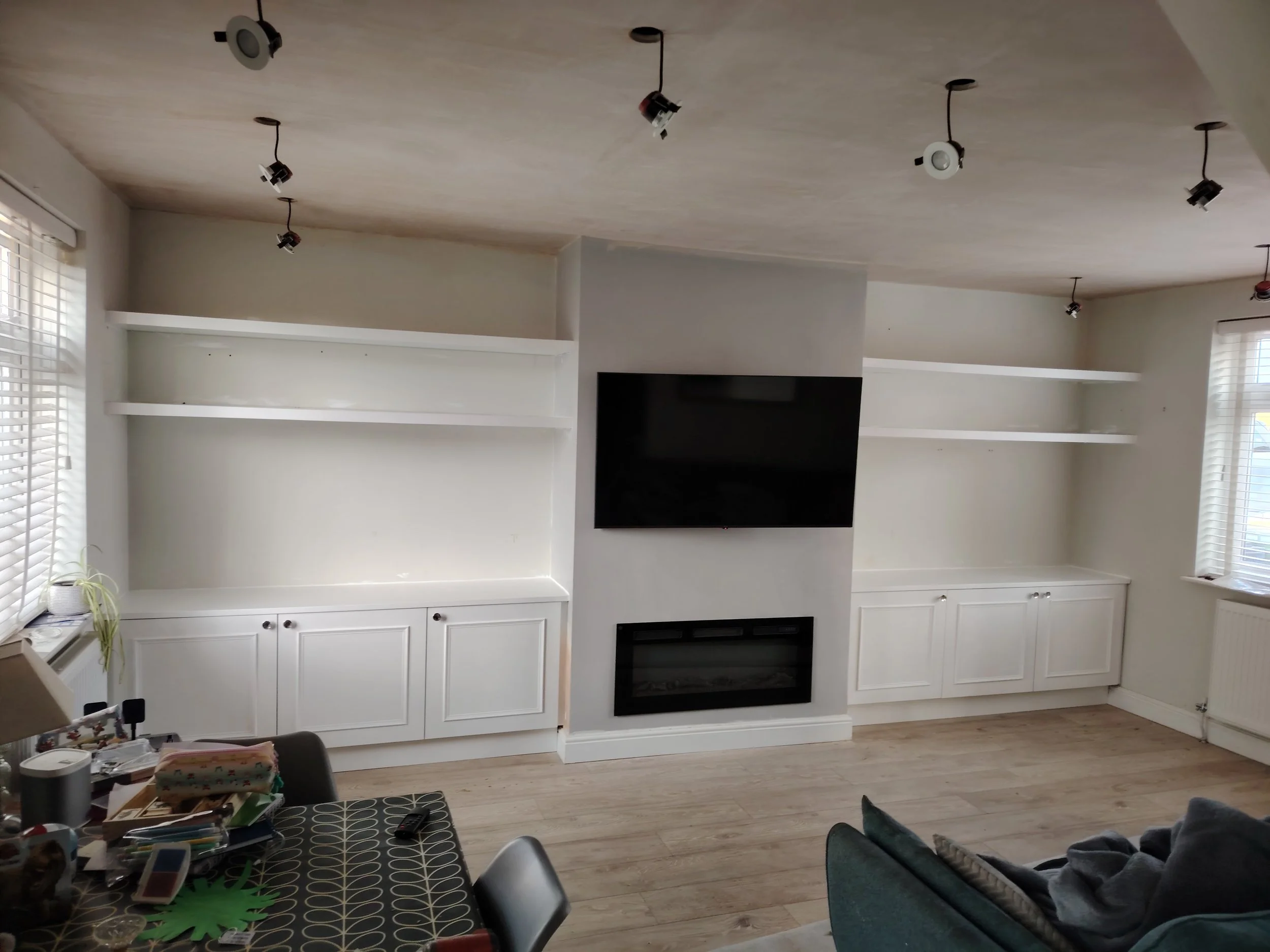 Living room with white built-in shelves, a flat-screen TV mounted on the wall, a fireplace below the TV, large windows with blinds, and a table with clutter in the foreground.