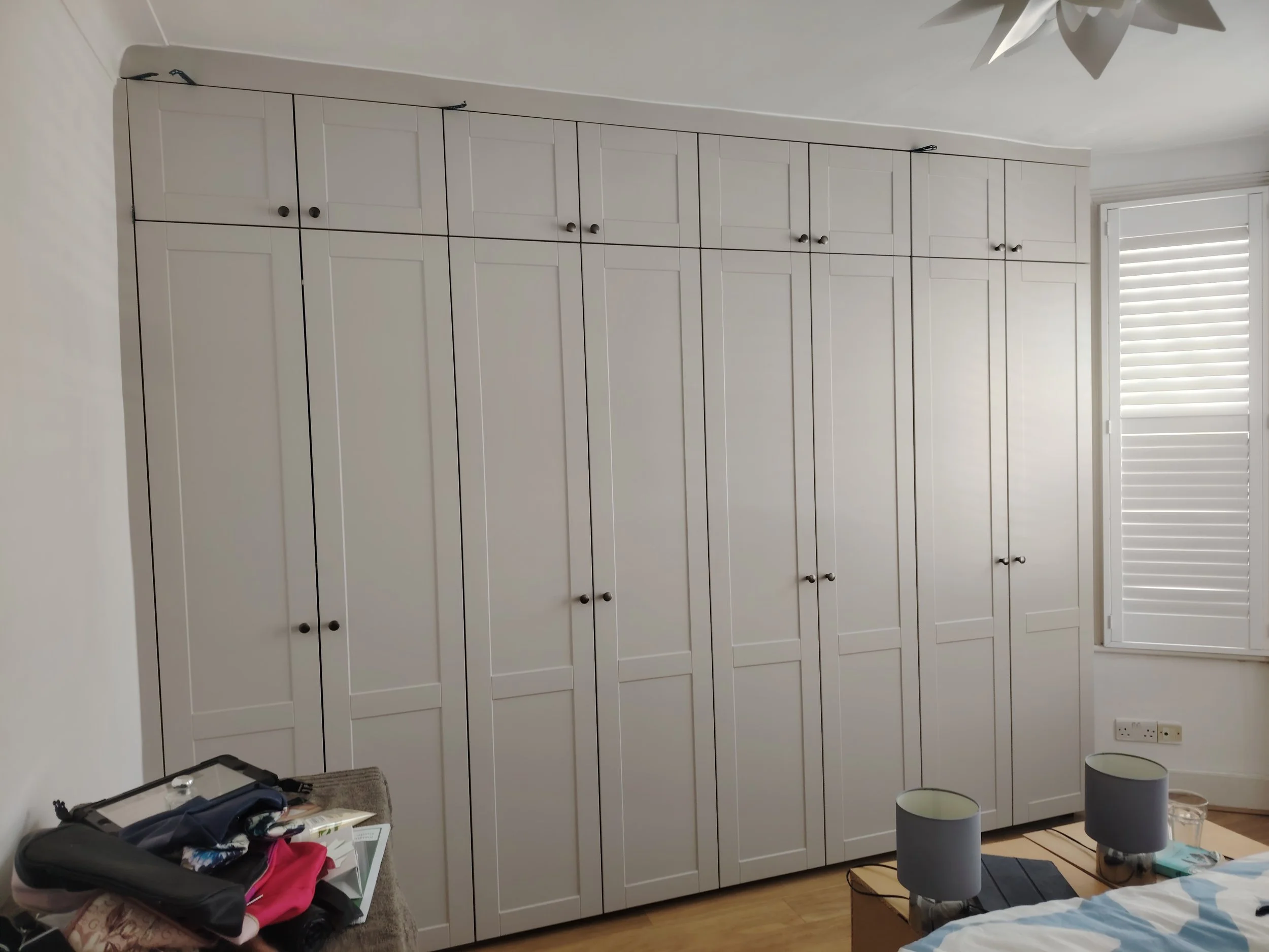 A large white built-in wardrobe with multiple doors in a bedroom. A window with blinds is on the right side. There is a nightstand with two lamps, clothes, and other items in the foreground.