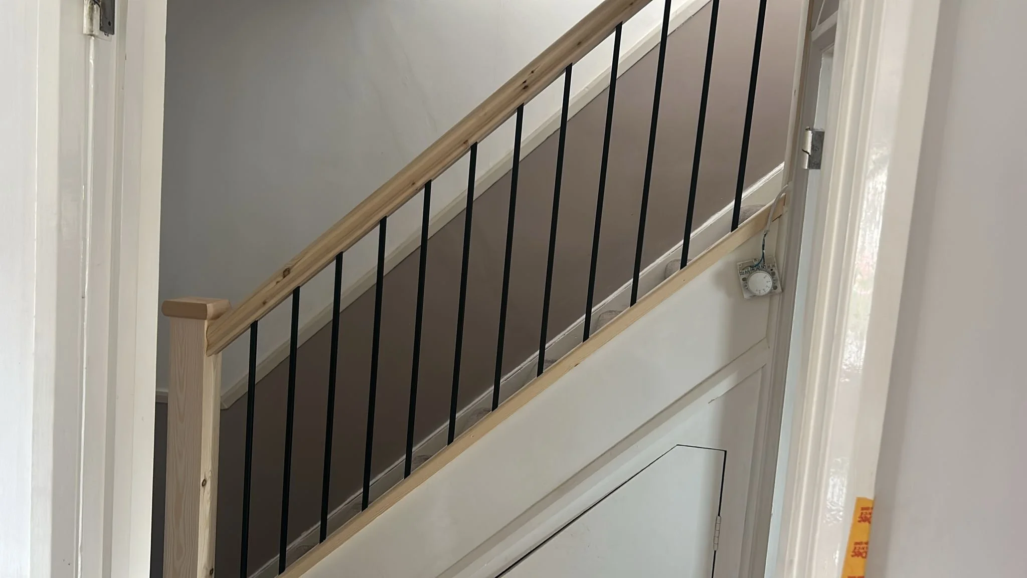 Part of an indoor staircase with exposed wooden handrail and black metal balusters, viewed through a doorway with white trim, with an unfinished wall and a white thermostat on the lower right side.