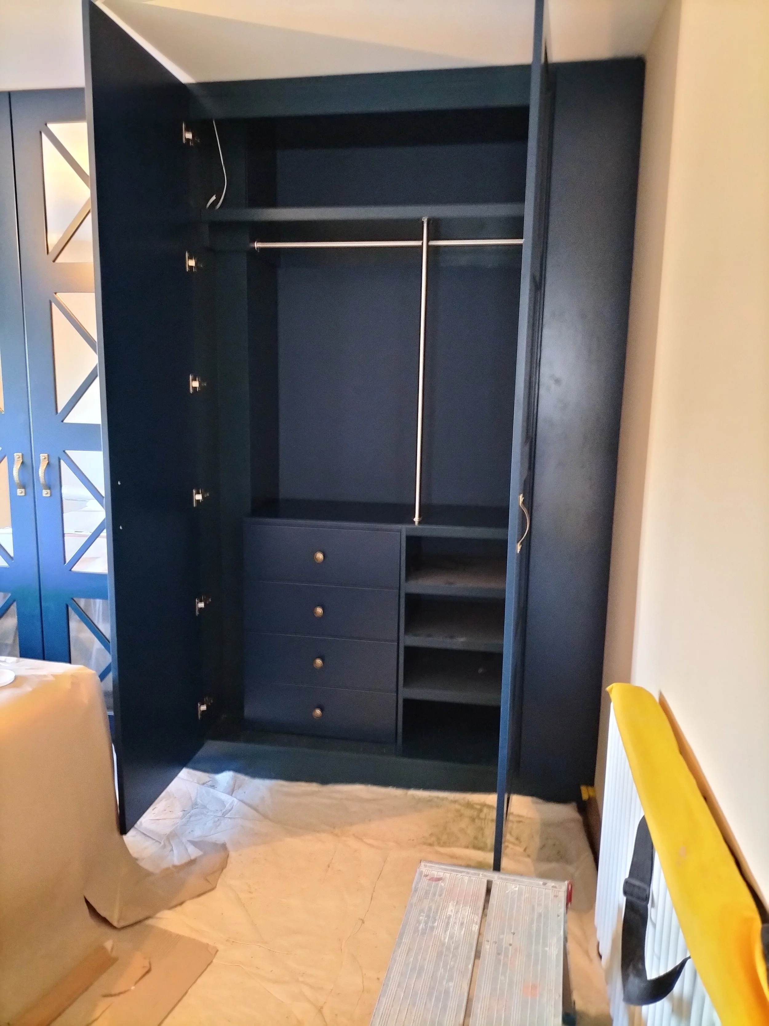 An open wardrobe with dark blue interior, metal hanging rod, three drawers, and shelves, in a room with beige walls and tarpaulin on the floor.