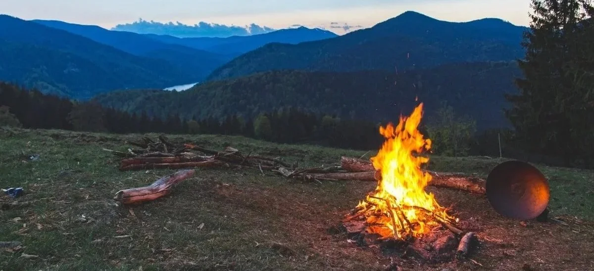 A campfire burning on a grassy area with logs and wood around it, overlooking a mountain landscape with a lake in the distance at dusk.