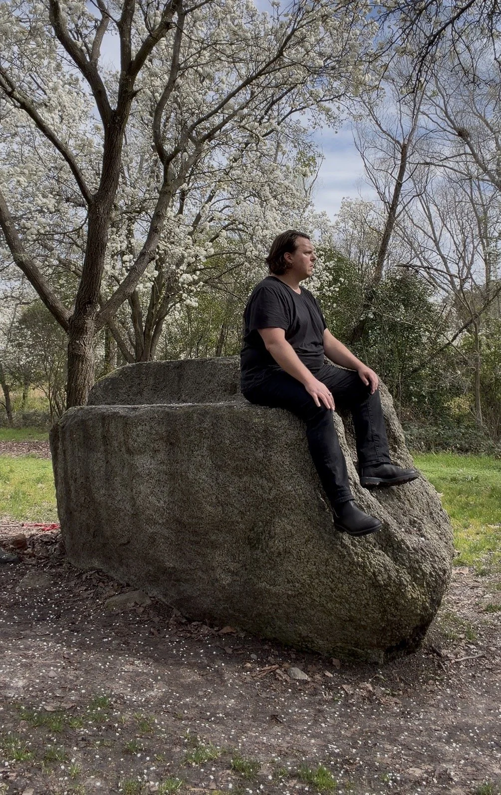 A man sitting on a large rock outdoors, with trees and a partly cloudy sky in the background.